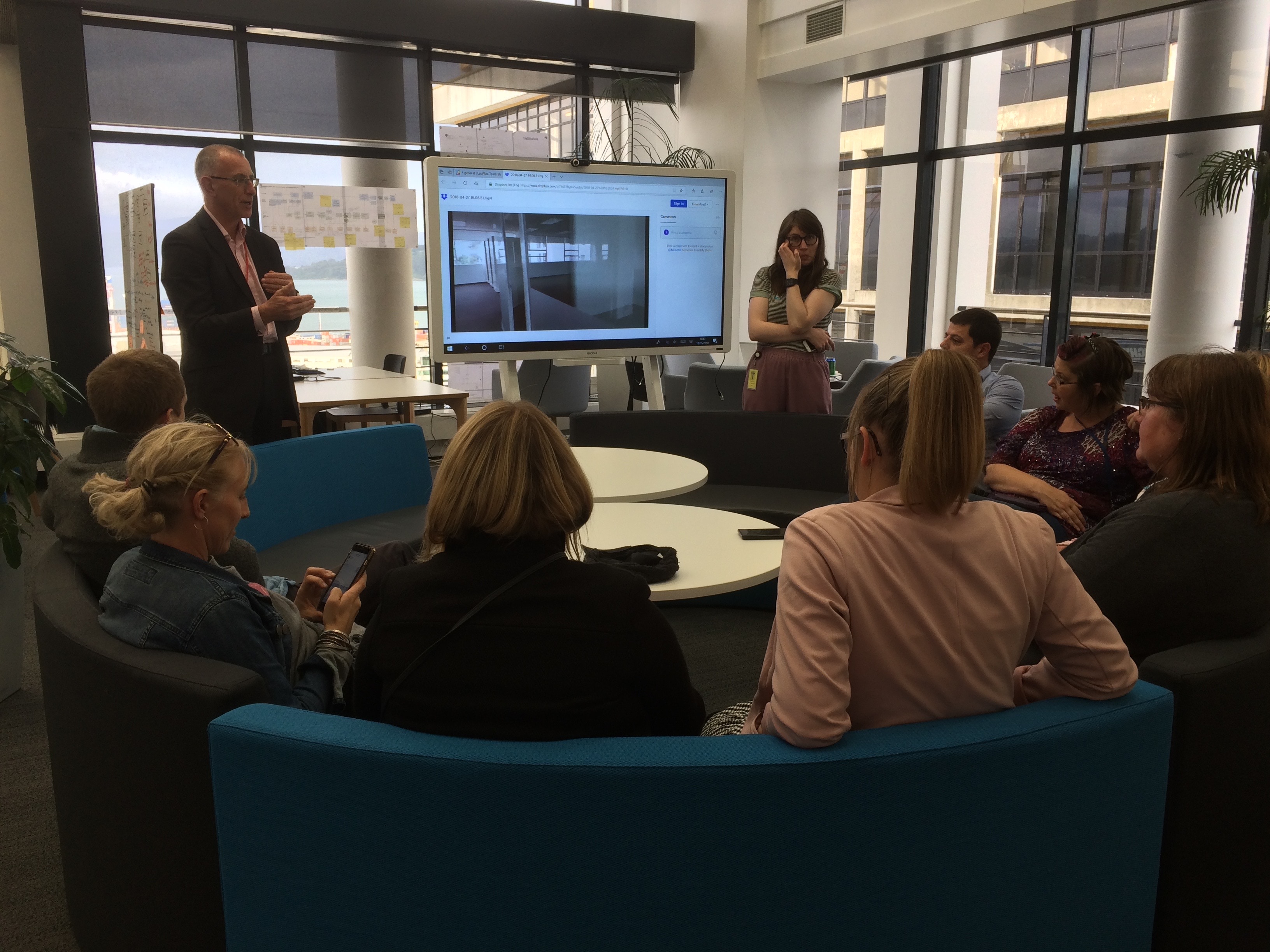 A gender- and age-diverse group sits in a circle in front of a large screen