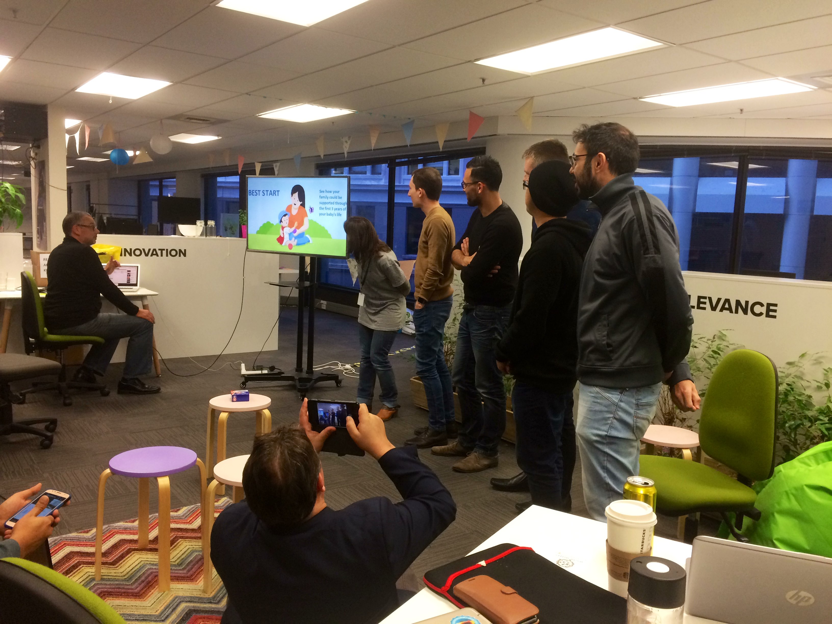 A group of people stand next to a screen in a colourful environment while an audience takes pictures of them